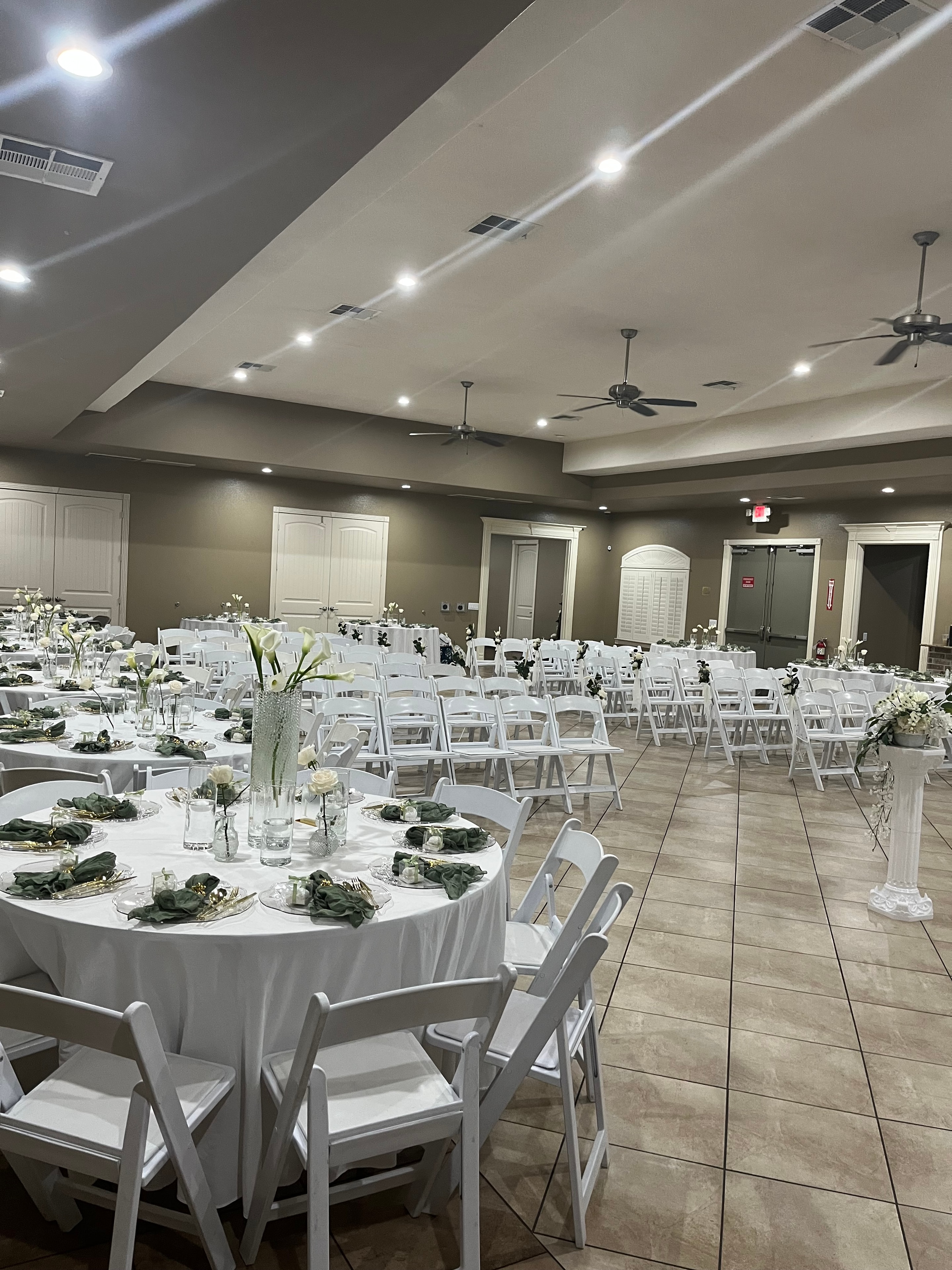 Wedding reception hall with white chairs, round tables, floral arrangements, and ceiling fans